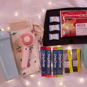 May include: An assortment of self-care items, including a light blue shaker bottle, floral socks, a pink handheld fan, and packets of DripDrop and LMNT. A black heat therapy pack and a small, light blue pill case are also present.