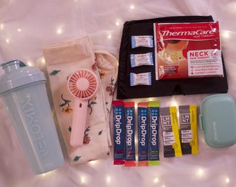 May include: An assortment of self-care items, including a light blue shaker bottle, floral socks, a pink handheld fan, and packets of DripDrop and LMNT. A black heat therapy pack and a small, light blue pill case are also present.