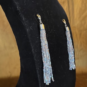 May include: A pair of drop earrings featuring long, beaded tassels. The tassels are a light blue hue with iridescent beads, and the earrings have silver-coloured metal findings. The earrings are displayed on a black velvet bust.