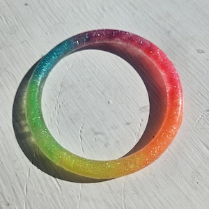 Puede incluir: Un brazalete de resina con los colores del arcoíris. El brazalete hace la transición del azul al verde, amarillo, naranja y rojo. La resina transparente está incrustada con purpurina, añadiendo brillo. El brazalete circular tiene aproximadamente 6,35 cm de diámetro.