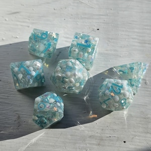 Puede incluir: Un conjunto de siete dados con resina transparente de color azul mar. Cada dado está incrustado con cuentas blancas, pequeños detalles azules y números. Los dados tienen varias formas geométricas, incluyendo cubos y un dado de múltiples caras.