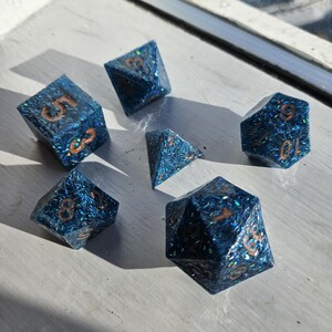 May include: A set of seven polyhedral dice in various shapes. The dice are a translucent blue with copper-colored numbers and glitter embedded throughout. The dice are arranged on a light gray surface.