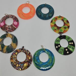 Puede incluir: Una colección de ocho colgantes circulares coloridos con un agujero en el centro. Los colgantes presentan varios diseños, incluyendo patrones florales, purpurina y remolinos abstractos. Cada colgante tiene un lazo plateado para colgar. Medidas en centímetros.