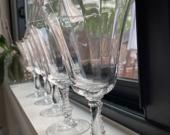 May include: A row of clear glass wine glasses with beaded stems. The glasses are arranged on a white windowsill.