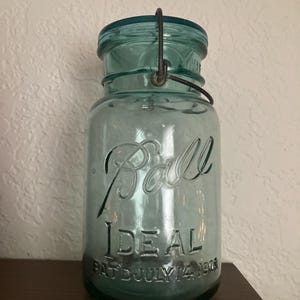 May include: A vintage blue-green Ball Ideal jar with a wire closure. The jar has the word "Ball" in script and "Ideal" embossed on the front, along with "Pat July 14 1908". The jar is made of glass and has a metal lid.