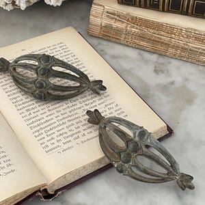 May include: Two antique metal decorative ornaments with an openwork design, resting on the pages of an old book. The ornaments have a tarnished, aged appearance, with a central cluster of small circles.