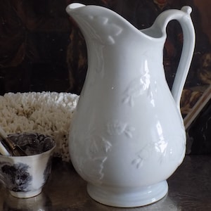 May include: White ceramic pitcher with a floral embossed design. The pitcher has a wide mouth and a handle.
