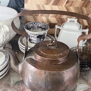 May include: A copper kettle with a large handle and lid, alongside a stack of plates, a teacup, and a sugar bowl. The kettle has a rounded body and a spout. The teacup has a black and white design. The items are on a decorative silver tray.