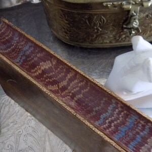 May include: The spine of a vintage book with a burgundy leather cover and gold gilded edges. The cover features a decorative pattern of blue and gold swirls.