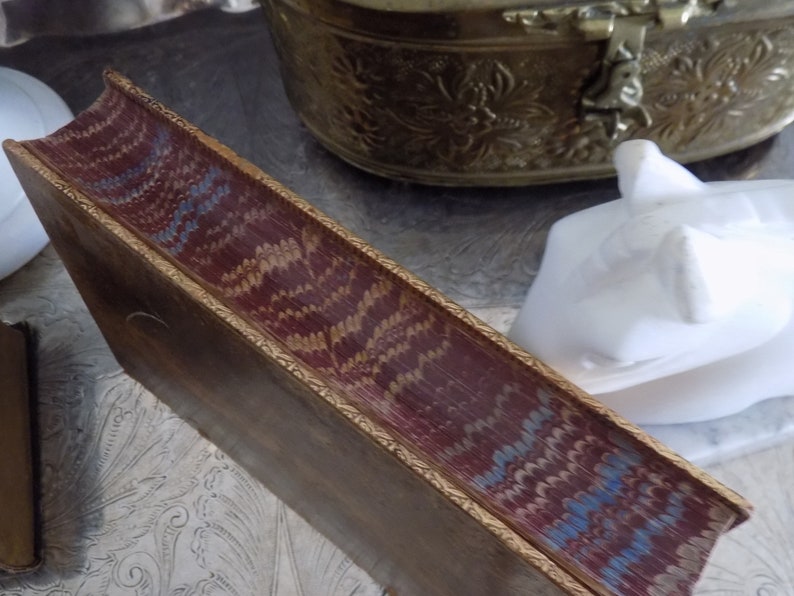 May include: The spine of a vintage book with a burgundy leather cover and gold gilded edges. The cover features a decorative pattern of blue and gold swirls.