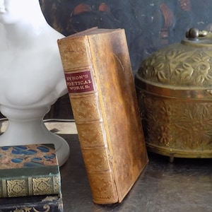 May include: A vintage, leather-bound book titled "Byron's Poetical Works." The book is brown with gold accents and is sitting on a table with other books and a decorative brass box.