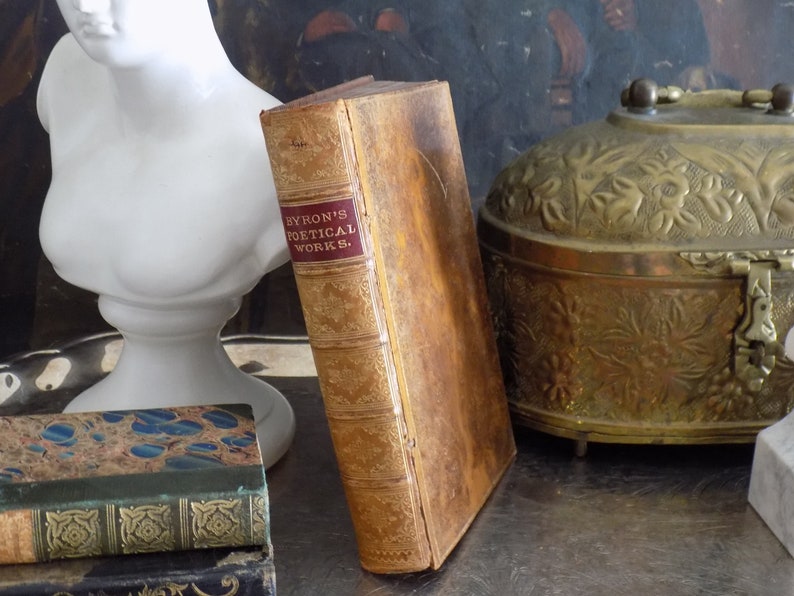 May include: A vintage, leather-bound book titled "Byron's Poetical Works." The book is brown with gold accents and is sitting on a table with other books and a decorative brass box.