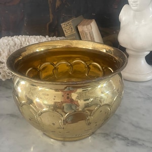 May include: A vintage, round brass planter with a scalloped design around the base. The planter has a wide, slightly flared rim and a polished, reflective surface. The interior is a warm, golden color. A decorative piece for home decor.