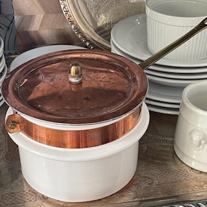 May include: A white ceramic pot with a copper band around the top and a matching copper lid featuring a small brass knob. A long, thin brass handle extends from the side of the pot, which is designed with a smooth, cylindrical body.
