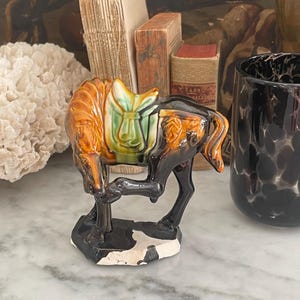 May include: A glazed ceramic horse figurine featuring a glossy brown mane and tail, a black body, and a light green saddle. The horse is depicted in a dynamic pose with one leg raised, standing on a black and white irregular base.