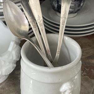 May include: A white ceramic cylindrical container with a small molded relief detail on the side, holding three silver-toned metal spoons. The spoons feature a slender handle design with a floral motif near the top and a smooth, rounded bowl.