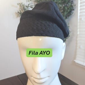 May include: A black beret hat made of textured fabric. The hat is displayed on a white mannequin head. The text "Fila AYO" is on a green rectangle on the mannequin. The beret is a classic style, suitable for various outfits.