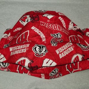 May include: Red face mask with a white pattern of Wisconsin Badgers logos, including the mascot Bucky Badger. The mask has two white elastic ear loops.
