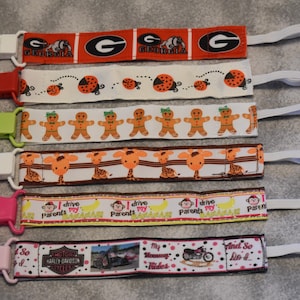 May include: Set of 7 colorful pacifier clips with various designs, including a University of Georgia logo, gingerbread men, giraffes, monkeys, and a Harley Davidson logo. Each clip has a white elastic strap.