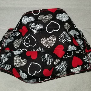 Puede incluir: Una mascarilla facial negra con un patrón de corazones rojos y blancos. La mascarilla tiene lazos elásticos para las orejas.