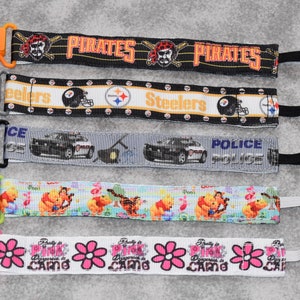 May include: Five colorful pacifier clips with different designs. The clips are made of plastic and have a variety of colors, including orange, gray, black, green, and pink. The designs include a pirate skull, a football helmet, a police car, Winnie the Pooh, and a pink flower.
