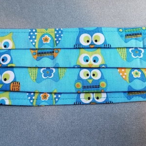 May include: A blue face mask with a repeating pattern of owls in various colors, including green, orange, and blue. The owls have large eyes and are set against a light blue background.