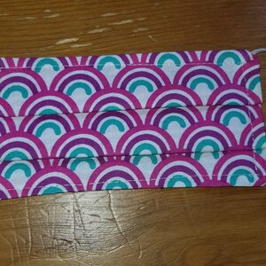 Puede incluir: Una mascarilla facial blanca con un patrón repetido de arcoíris turquesa, morado y rosa. La mascarilla tiene lazos elásticos para las orejas.