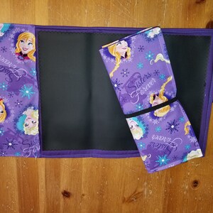May include: A purple fabric roll-up mat with a black center and a Frozen theme. The fabric features images of Elsa and Anna, snowflakes, and the text "Sisters Forever".