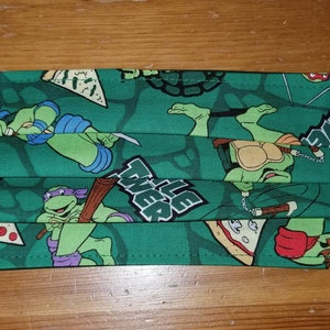 Op de afbeelding: Groen stoffen mondkapje met een Teenage Mutant Ninja Turtles-print. Het masker heeft afbeeldingen van de turtles, pizzapunten en de tekst "Teenage Mutant Ninja Turtles".
