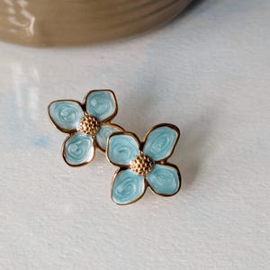 Puede incluir: Un par de pendientes de botón con forma de flor. Cada pendiente presenta cuatro pétalos de color azul claro con bordes dorados y un centro dorado texturizado. Los pendientes están sobre un fondo suave y claro.