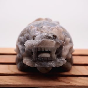 May include: A detailed, carved stone sculpture of a mythical creature with an open mouth and visible teeth. The sculpture is primarily gray and beige, with intricate details. The background is a plain white, and the sculpture rests on a wooden surface.