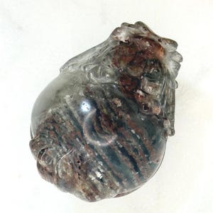 May include: A carved stone sculpture with a clear, rounded top and a body featuring streaks of brown, red, and gray. The sculpture has a detailed, textured surface and appears to be a decorative art piece.