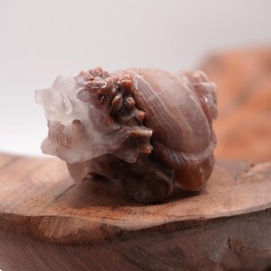 May include: A carved stone sculpture of a mythical creature, possibly a dragon or griffin, in shades of brown and white. The sculpture rests on a wooden base with a natural grain pattern. The artwork is detailed and appears to be hand-carved.