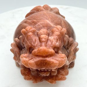 Puede incluir: Una escultura de piedra tallada de color marrón rojizo de una criatura mítica. La escultura presenta rasgos faciales detallados, incluyendo un hocico prominente, dientes y una melena estilizada. El diseño sugiere un dragón o bestia mitológica similar.