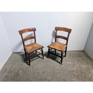 Op de afbeelding: Twee antieke houten stoelen met geweven zittingen. De stoelen hebben een donkerbruine afwerking en een eenvoudig, klassiek ontwerp. De zittingen lijken gemaakt te zijn van geweven stro of een vergelijkbaar materiaal. De stoelen staan naast elkaar tegen een effen witte muur.