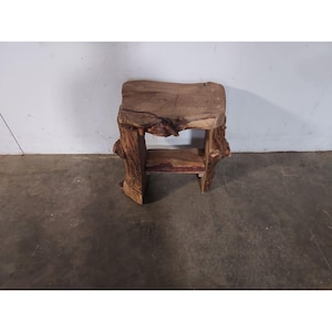 May include: A rustic wooden stool with a natural wood finish. The stool has a rectangular top and a unique, irregular base made from a tree trunk.