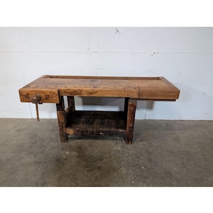 Puede incluir: Un banco de trabajo de madera vintage con un aspecto oscuro y envejecido. El banco de trabajo cuenta con un tornillo de banco incorporado en el lado izquierdo y un estante inferior. La madera tiene vetas y nudos visibles, mostrando su carácter rústico. El banco de trabajo está soportado por patas robustas.