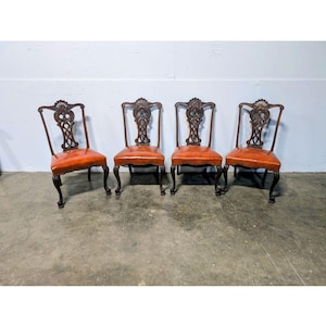 Op de afbeelding: Een set van vier stoelen van donker hout met oranje lederen zittingen en ingewikkeld gesneden rugleuningen met opengewerkte details. Elke stoel staat op vier gebogen poten en heeft een decoratief schelpachtig motief bovenaan de rugleuning.