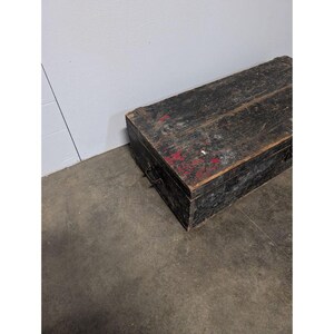 May include: A weathered, rectangular wooden chest painted black with red accents. The chest has a hinged lid and metal handles on the sides. The wood grain is visible, and the chest appears aged. It sits on a concrete floor.
