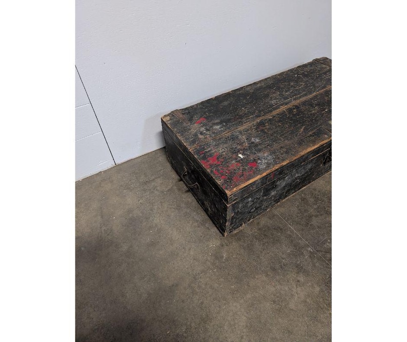 May include: A weathered, rectangular wooden chest painted black with red accents. The chest has a hinged lid and metal handles on the sides. The wood grain is visible, and the chest appears aged. It sits on a concrete floor.