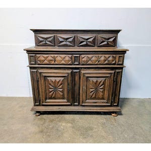 Può includere: Un mobile in legno marrone scuro con intricati disegni geometrici intagliati. Il mobile presenta due ante con motivi a stella, due cassetti e un piano decorativo. Il pezzo ha un'estetica vintage e poggia su piccoli piedi arrotondati.