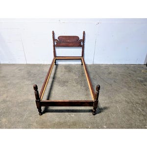 May include: A vintage wooden bed frame with a headboard and footboard. The frame is a dark brown color and appears to be made of solid wood. The headboard has a decorative top. The bed frame is designed for a twin-sized mattress.