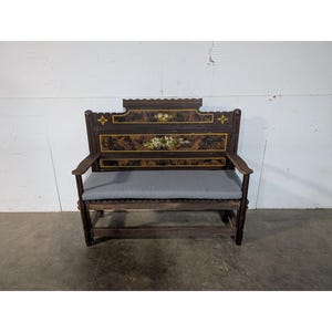 May include: A dark brown wooden bench with a gray cushion. The back of the bench has a decorative design with painted flowers and gold accents.