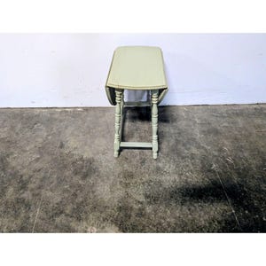 May include: A light sage green drop-leaf table featuring a rectangular top with rounded edges and four turned, spindle-style legs connected by horizontal stretchers. The table is shown from a front-facing perspective with both leaves folded down.