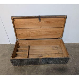 May include: An open, rectangular wooden chest with a hinged lid. The chest is made of light-colored wood with a dark, weathered exterior. A small, stepped compartment is visible inside. The lid is secured with a metal clasp.