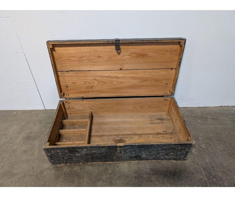 May include: An open, rectangular wooden chest with a hinged lid. The chest is made of light-colored wood with a dark, weathered exterior. A small, stepped compartment is visible inside. The lid is secured with a metal clasp.
