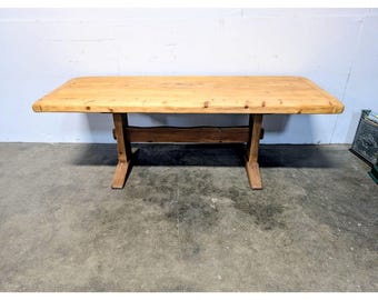Puede incluir: Una mesa rectangular de madera con un tablero de tono claro y superficie lisa con esquinas redondeadas. La base consta de dos soportes verticales conectados por un travesaño horizontal, todo en un tono de madera oscuro.