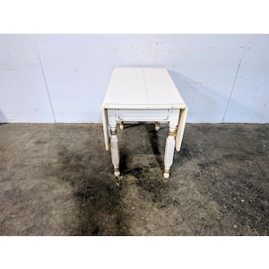 Pode incluir: Uma mesa branca retangular com tampo rebatível e quatro pernas ornamentadas. A mesa tem uma gaveta e detalhes dourados nas pernas. A mesa tem aproximadamente 76 cm de comprimento quando as folhas estão abaixadas.