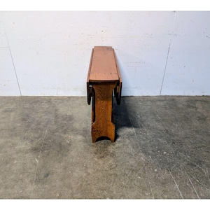Puede incluir: Una mesa plegable de madera vintage con un acabado marrón rojizo cálido. La mesa tiene una tapa rectangular y una base decorativa con detalles tallados. Las hojas abatibles están plegadas y la mesa mide unos 76 cm de largo.