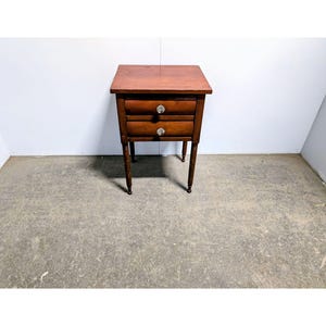 May include: A brown wooden nightstand featuring a rectangular top, two stacked drawers with clear glass knobs, and four slender, turned legs. The piece has a smooth, polished finish and a simple, functional design with clean lines and a warm tone.